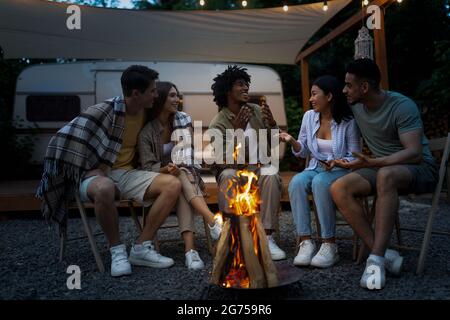 Multiethnische Freunde sitzen am Lagerfeuer, unterhalten sich und verbringen gemeinsam Zeit auf einem Campingausflug mit dem Wohnmobil Stockfoto