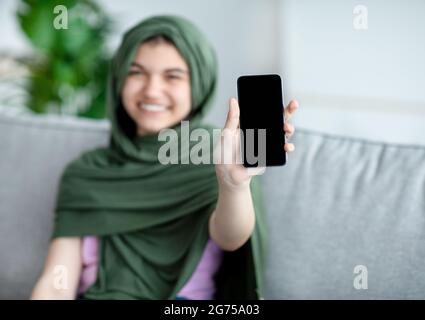 Muslimische Teenager-Mädchen in Hijab zeigt Handy mit leerem Bildschirm zu Hause, Mockup für mobile App oder Website-Design Stockfoto