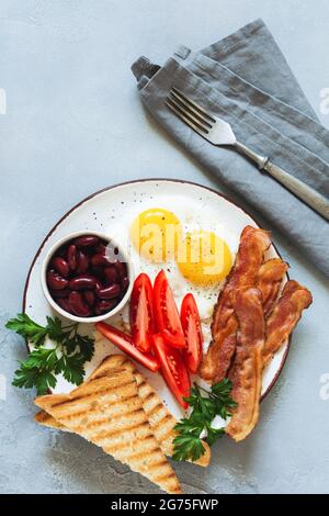 Traditionelles englisches Frühstück mit Spiegeleiern, Würstchen, Bohnen, gegrillten Tomaten und Speck auf grauem Betongrund. Draufsicht Stockfoto