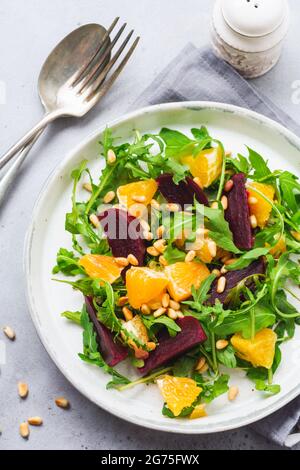 Salat aus Rucola, gebackene Rote Bete, Orangen- und rote Zwiebeln mit Gewürzen und Pinienkernen in einer weißen Schale. Olivenöl und Zutaten auf dem Tisch aus grauem Stein. Auswahl Stockfoto