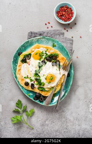 Türkische Eier Fladenbrot mit Joghurt, Käse, Oliven, Spinat und rotem Pfeffer auf Keramik-Vintage-Teller auf grauem altem Hintergrund. Draufsicht. Stockfoto