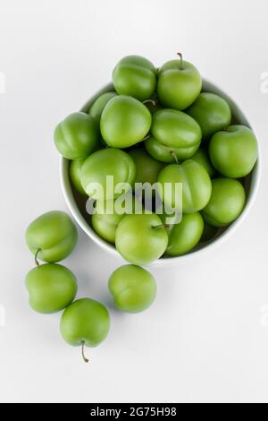 Eine vertikale Draufsicht auf einen Haufen saurer grüner Pflaumen in einer Schüssel auf einem schlichten weißen Hintergrund Stockfoto