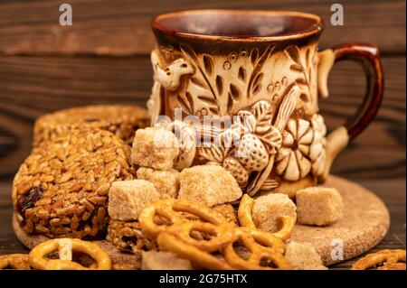 Hausgemachte Kekse mit Sonnenblumenkernen und Rosinen, Stücke von braunem Rohrzucker, geformten Brotstäbchen und einem Becher Tee aus Steingut auf einem Holztisch. Clo Stockfoto