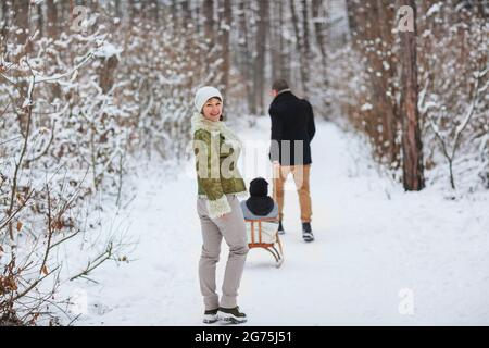 Fröhliche Eltern, die hinter dem fröhlichen Jungen mit Schlitten herumlaufen, während sie am Wintertag Zeit in verschneiten Wäldern verbringen Stockfoto