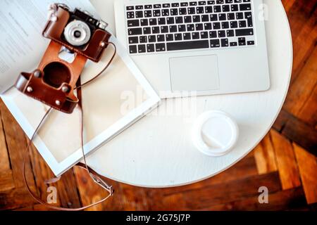 Retro-Fotokamera und Tasse Kaffee, um auf dem runden Tisch in der Nähe von geöffnetem Magazin und Netbook mit leerem Bildschirm im kreativen Studio zu gehen