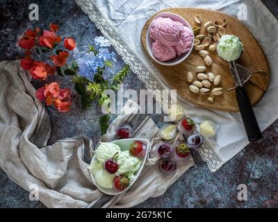 Schüssel und Kugeln mit köstlichem Himbeer-, Kirsch- und Pistazien-Eis.Holzteller mit köstlichem Eis und verschiedenen Zutaten auf dem Tisch Stockfoto