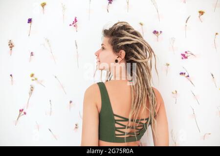 Seitenansicht der jungen Sportlerin in Sportkleidung mit Dreadlocks und Blick auf die Kamera vor weißem Hintergrund mit zarten Blumen nach dem Training Stockfoto