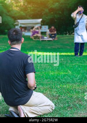 Rückansicht des asiatischen Mannes mit Kamera auf Gras sitzend, der sich vorbereitete, mit seiner Familie im Garten Fotos zu machen. Glückliche Familie, die draußen Zeit zusammen verbringt Stockfoto