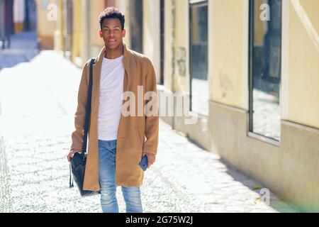 Ein junger schwarzer Mann, der die Straße entlang läuft, trägt eine Aktentasche und ein Smartphone. Stockfoto