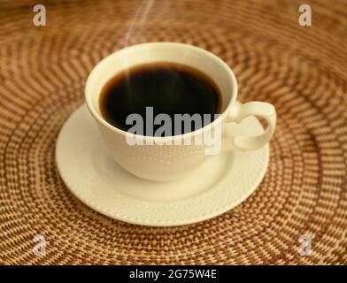 Ein weißer Becher mit dampfendem schwarzen Kaffee auf einer braun gewebten Tischdecke Stockfoto