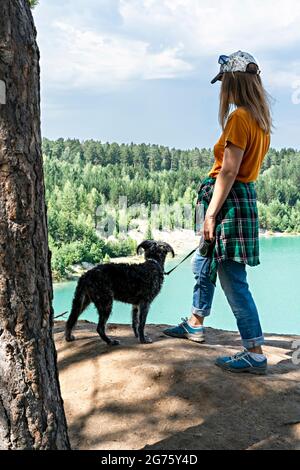 Rückansicht einer jungen blonden Frau, die in einer Mütze und einem Ingwer-T-Shirt auf einer Klippe mit einem blau-türkisfarbenen See oder Fluss und einem Nadelwaldmit flauschigen d steht Stockfoto