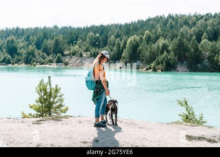 Rückansicht einer jungen Frau, die in einer Mütze mit Rucksack unterwegs ist und am sandigen Ufer des blauen Sees oder des Flusses mit einem flauschigen grauen Hund läuft, Haustier liebt aktiven Lebensstil, t Stockfoto