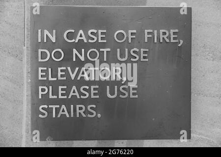 Ein Schild mit der Aufschrift: Im Brandfall keinen Aufzug verwenden, bitte Treppen verwenden, die die Mitarbeiter darauf verweisen, was sie im Brandfall tun sollen. Schwarz und W Stockfoto