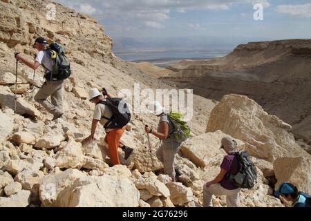 Ältere Wanderer, ältere Erwachsene, erklimmen zwischen Felsbrocken die steilen Hänge oberhalb von Nachal Himrit, der judäischen Wüste mit Blick auf das Tote Meer und Herbstwolken Stockfoto