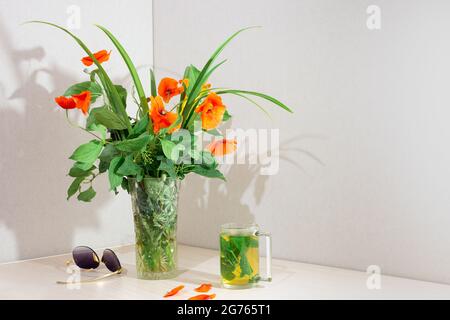 Ein Strauß Mohnblumen in einer Glasvase auf dem Tisch im Zimmer. In der Nähe befindet sich eine Minze Minztee und Sonnenbrille. Innenecke mit Kopierraum. Stockfoto