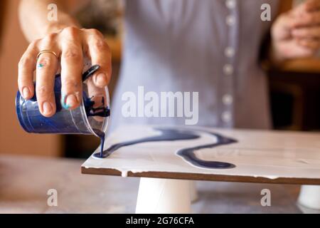Frau mit der rechten Hand gießt blaue Farbe aus dem Becher auf Leinwand. .Master Klasse. Kreativität und Design. Künstler bei der Arbeit. Freiheit und Inspiration Stockfoto