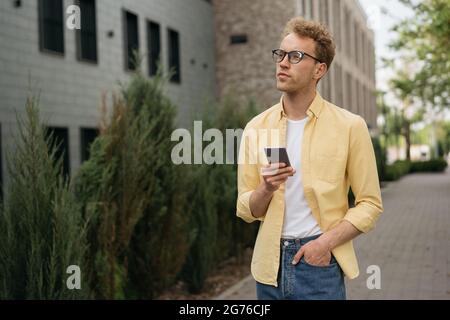 Schöner nachdenklicher Mann in gelbem Casual-Hemd und stilvoller Brille mit Mobiltelefon, der auf der Straße unterwegs ist, um Platz zu kopieren Stockfoto