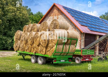 Grüner Anhänger gefüllt mit Heuballen, die vor der Ziegelsteinscheune auf einem Bauernhof geparkt sind. Solarpaneele auf dem Dach der Scheune installiert. Stockfoto