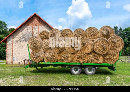 Grüner Anhänger gefüllt mit Heuballen, die vor der Ziegelsteinscheune auf einem Bauernhof geparkt sind. Stockfoto