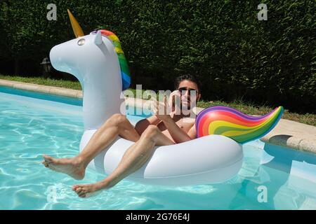 Digitaler Nomade in Sonnenbrillen, der auf einem aufblasbaren Einhorn-Ring in einem Schwimmbad rückwärts am Telefon spricht. Sommerkonzept. Stockfoto