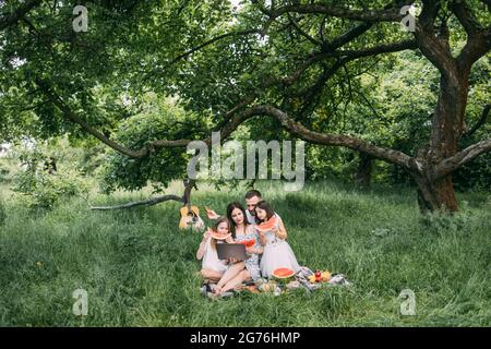 Junge Eltern mit zwei kleinen Töchtern, die während des Picknicks im grünen Garten einen Laptop benutzen. Glückliche Familie, die ihre Zeit im Freien genießt. Konzept von Mensch und Technik. Stockfoto