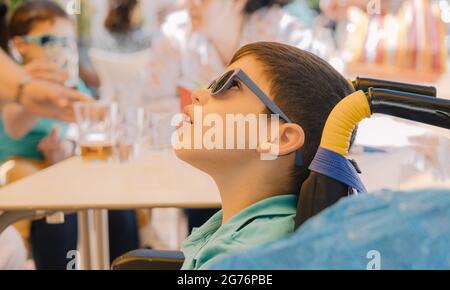 Junge mit Sonnenbrille sitzt in einem Rollstuhl aufgrund einer motorischen Behinderung seines Körpers an einem sonnigen Tag Stockfoto