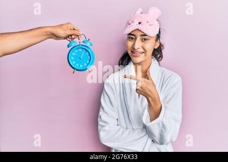 Hispanische Teenager-Mädchen mit Zahnspangen tragen Schlafmaske und Robe mit Wecker in der Nähe ihres lächelnden fröhlich zeigen mit Hand und Finger nach oben Stockfoto