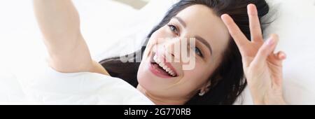 Porträt einer lächelnden jungen Frau auf weißem Bett, die Selfie nimmt Auf dem Smartphone Stockfoto
