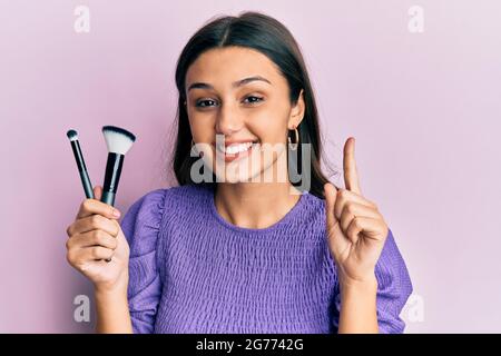 Junge hispanische Frau hält Make-up Pinsel lächeln mit einer Idee oder Frage zeigen Finger mit glücklichen Gesicht, Nummer eins Stockfoto