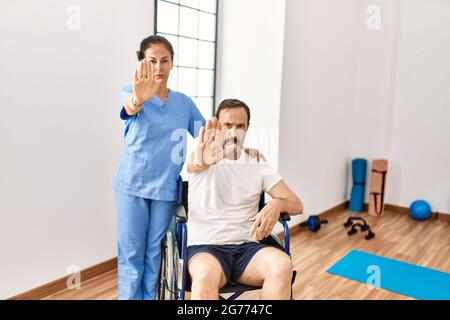 Hispanischer Mann mittleren Alters, der auf dem Rollstuhl sitzt und in der Rehabilitationsklinik mit offener Hand ein Stoppschild mit ernstem und selbstbewusster Ausdruck macht, Stockfoto