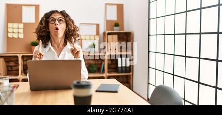 Eine hispanische Frau mittleren Alters, die im Büro mit einer Brille arbeitete, war erstaunt und überrascht, als sie nach oben schaute und mit Fingern und erhobenen Armen zeigte. Stockfoto
