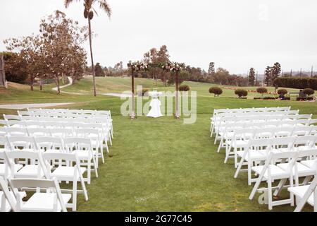 Hochzeitszeremonie im Freien an einem bewölkten Regentag - quadratischer Holzbogen mit Blumenarrangements, weiße Stühle auf grünem Rasen. Stockfoto