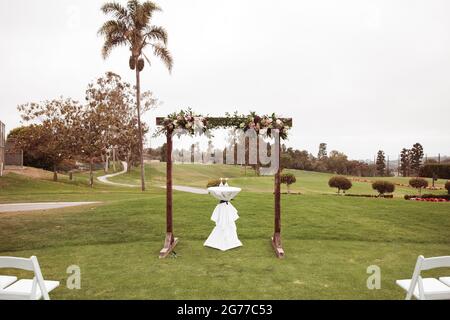 Hochzeitszeremonie im Freien an einem bewölkten Regentag - quadratischer Holzbogen mit Blumenarrangements, weiße Stühle auf grünem Rasen. Stockfoto