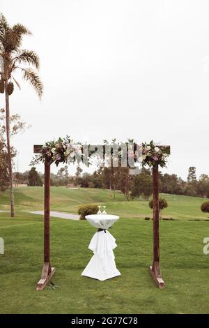 Hochzeitszeremonie im Freien an einem bewölkten Regentag - quadratischer Holzbogen mit Blumenarrangements, weiße Stühle auf grünem Rasen. Stockfoto