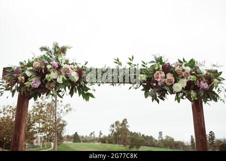 Hochzeitszeremonie im Freien an einem bewölkten Regentag - quadratischer Holzbogen mit Blumenarrangements, weiße Stühle auf grünem Rasen. Stockfoto