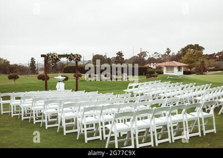 Hochzeitszeremonie im Freien an einem bewölkten Regentag - quadratischer Holzbogen mit Blumenarrangements, weiße Stühle auf grünem Rasen. Stockfoto