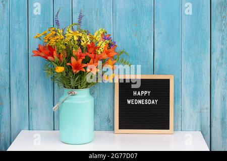 Happy Wednesday Worte auf schwarzem Briefbrett und Strauß von hellen Wildblumen in Dose Vase auf Tisch gegen blaue Holzwand. Konzept Hallo Mittwoch Stockfoto