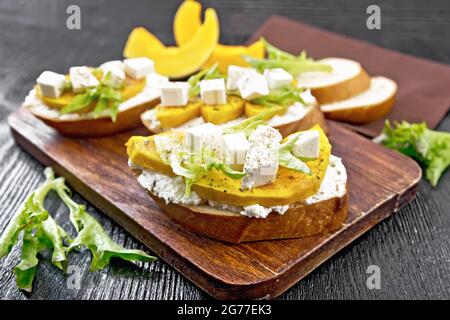 Bruschetta mit gebackenem Kürbis, gesalzenem Feta-Käse, Ricotta, Rucola und Gewürzen, Handtuch- und Gemüsescheiben auf Holzbrett-Hintergrund Stockfoto