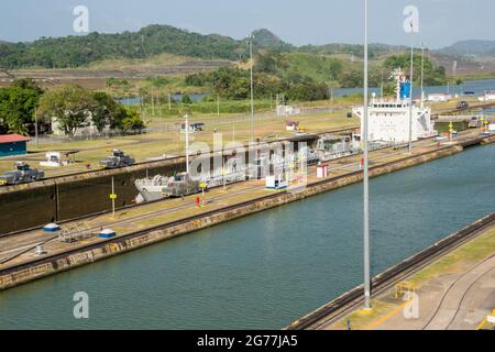 Eine große Ladung fuhr gerade an einem sonnigen Tag durch den Panamakanal. Die Hauptbüros und eine der Schleusen sind zu sehen. Stockfoto