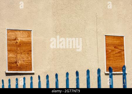 Zwei kleine Fenster mit Sperrholzplatten. Unscharfer Metallzaun im Vordergrund Stockfoto