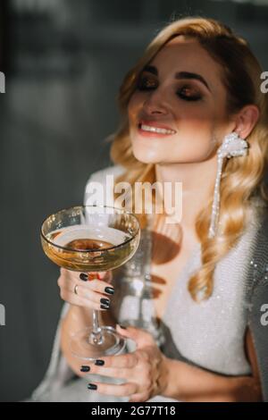 Junge schöne luxuriöse blonde Frau mit einem Glas in ihr Hands feiert das neue Jahr vor dem Hintergrund von Unschärfe Lichter Bokeh von Weihnachten Stockfoto