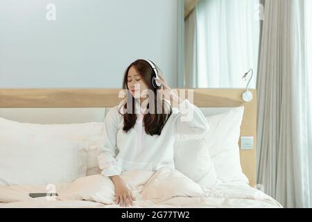 Hübsche junge Frau, die ihre Lieblingsmusik über Kopfhörer hört, wenn sie auf dem Bett sitzt Stockfoto