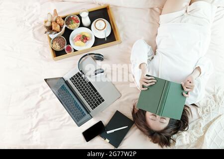 Faule junge Frau, die auf dem Bett mit geöffnetem Laptop und Tablett mit Frühstück lag und schlief, nachdem sie ein langweiliges Buch gelesen hatte Stockfoto