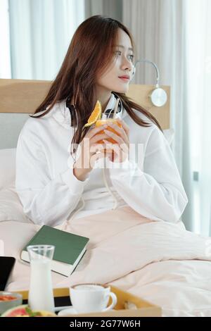 Porträt einer nachdenklichen jungen Frau, die im Bett sitzt und zum Frühstück frischen Orangensaft trinkt Stockfoto