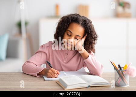 Lernkonzept. Müde und gelangweilte afroamerikanische Mädchen, die zu Hause am Schreibtisch sitzt, im Notizbuch schreibt, Hausaufgaben macht Stockfoto