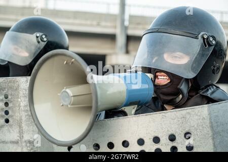 Polizist im Sturzhelm, der Schild hält und über Lautsprecher Nachrichten sendet, während er mit Terroristen von Demonstranten spricht Stockfoto