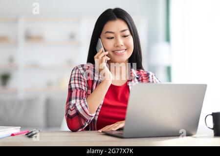 Telearbeitskonzept. Junge Koreanische Frau Im Gespräch Auf Handy Und Mit Laptop Stockfoto