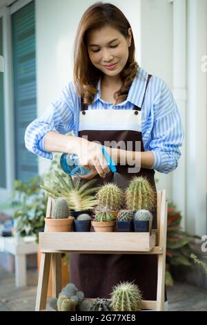 Nette asiatische Hausfrau trägt Schürze hält Wassersprüher und Bewässerung in den Topf von kleinen Pflanzen und Kaktus mit Lächeln Gesicht und glücklich mit Hobby an ho Stockfoto