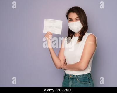 Frau trägt eine medizinische Schutzmaske mit Pflaster auf den Armhalten und zeigt eine geimpfte Karteikarte, nachdem sie den Impfstoff gegen Coronav erhalten hat Stockfoto