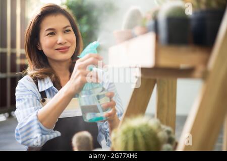Erwachsene asiatische niedliche 45er Frau trägt Schürze hält Sprayer Krug und sprühen Tröpfchen Wasser zu Kaktus im Regal mit glücklichem Lächeln Gesicht zu Hause. Entspannen Sie sich Stockfoto